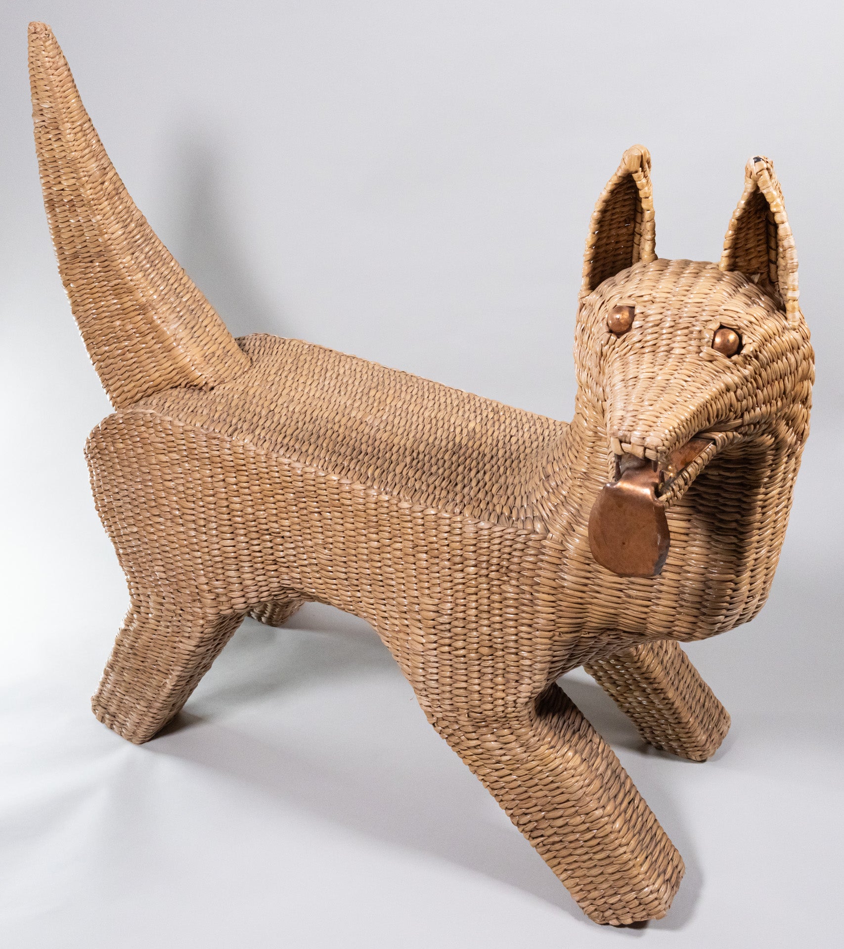 A seagrass woven over wire piece in the shape of a dog, looking to it's side with metal tongue hanging out.