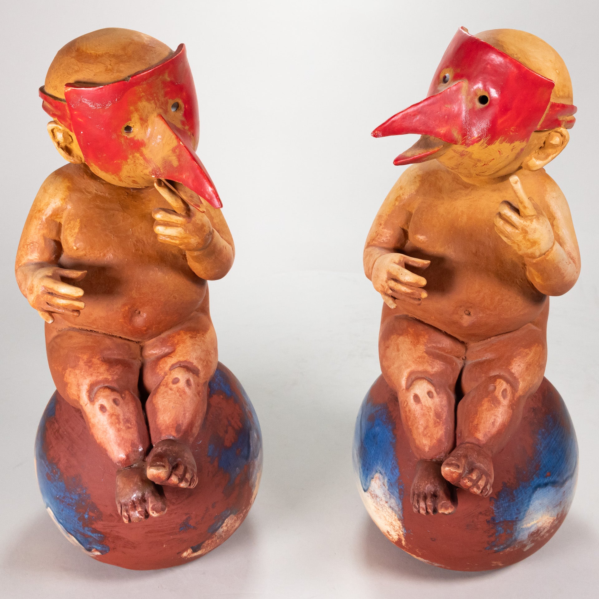 A pair of matching ceramic pieces of chubby babies wearing red masks with long beaks, both sitting on orbs.