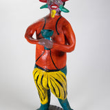 A tall ceramic sculpture of a devil holding a green cup and colorful horns around his face, and wearing an orange shirt with yellow and green bottoms.