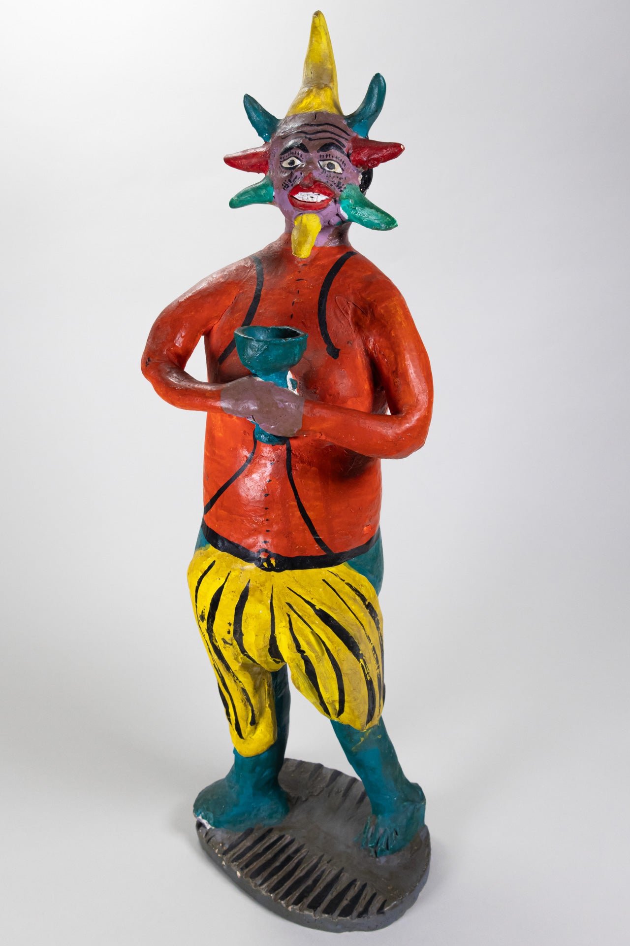 A tall ceramic sculpture of a devil holding a green cup and colorful horns around his face, and wearing an orange shirt with yellow and green bottoms.