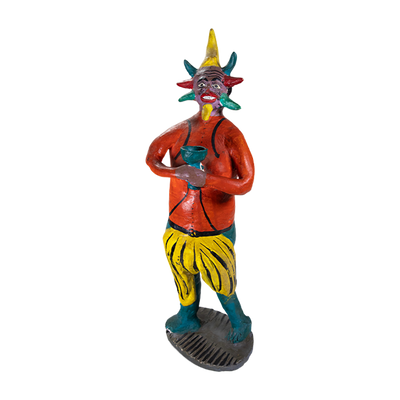 A tall ceramic sculpture of a devil holding a green cup and colorful horns around his face, and wearing an orange shirt with yellow and green bottoms.