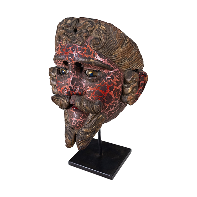 Red and gold painted wooden mask with facial hair and glasses, mounted on a metal stand.