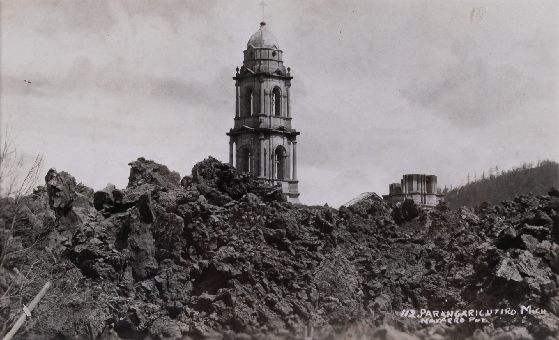 Black and white postcard of the Church of San Juan Parangaricutiro surrounded by the ash from the aftermath of  the eruption of Parícutin.
