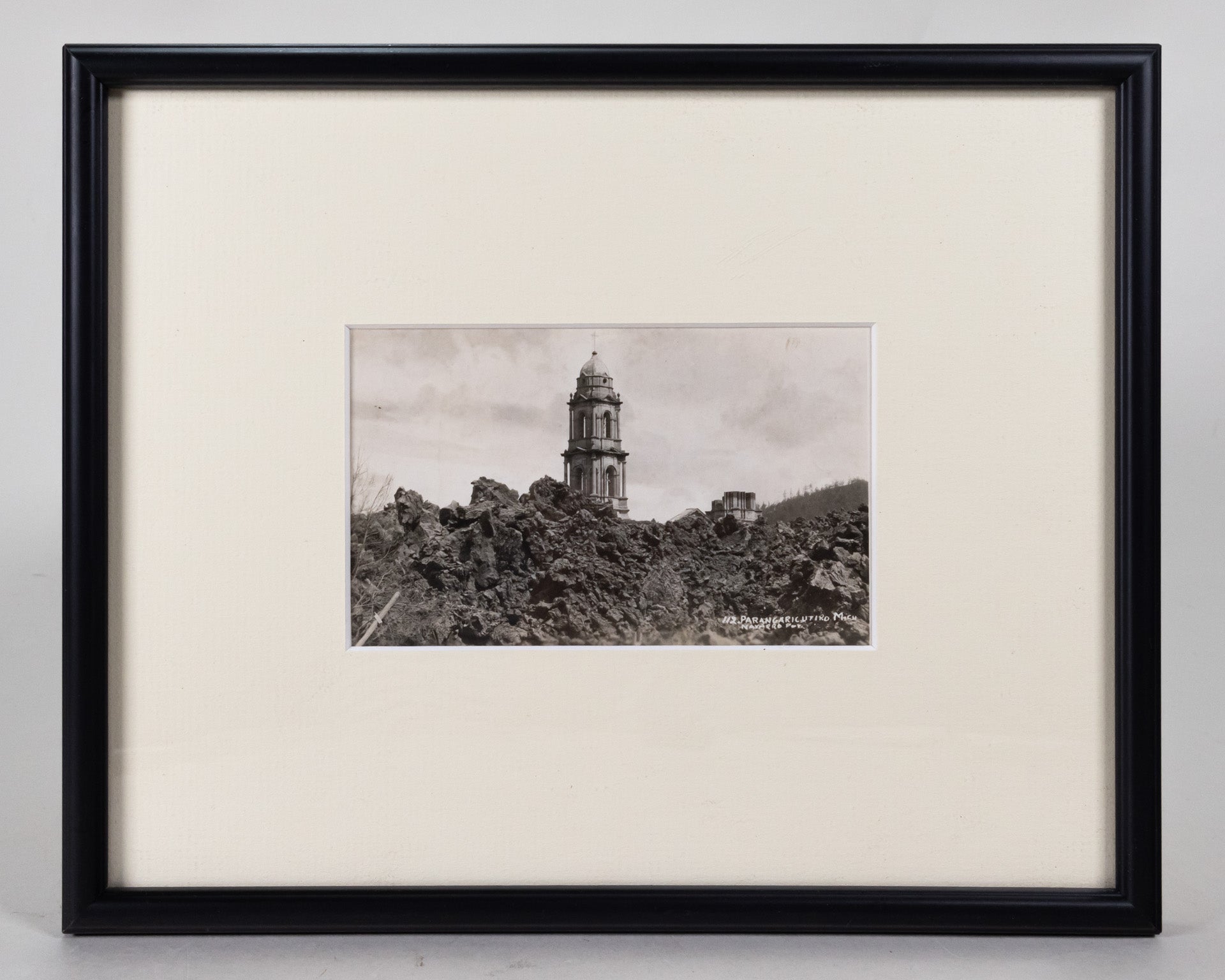 Framed black and white postcard of the Church of San Juan Parangaricutiro surrounded by the ash from the aftermath of  the eruption of Parícutin.