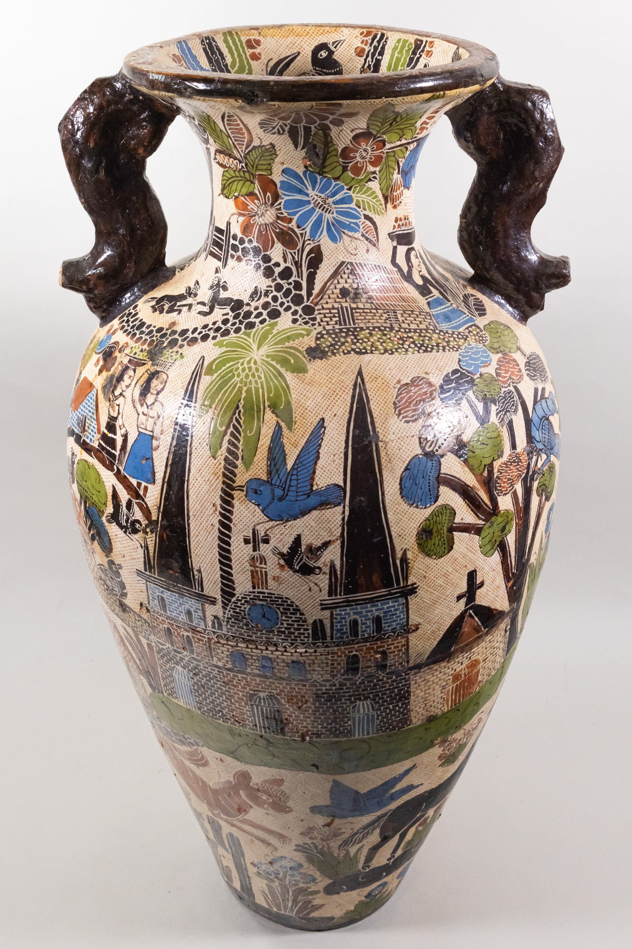 Large ceramic beige vase with crosshatches and brown handles, with elaborate artwork featuring church buildings, houses, villagers and animals.
